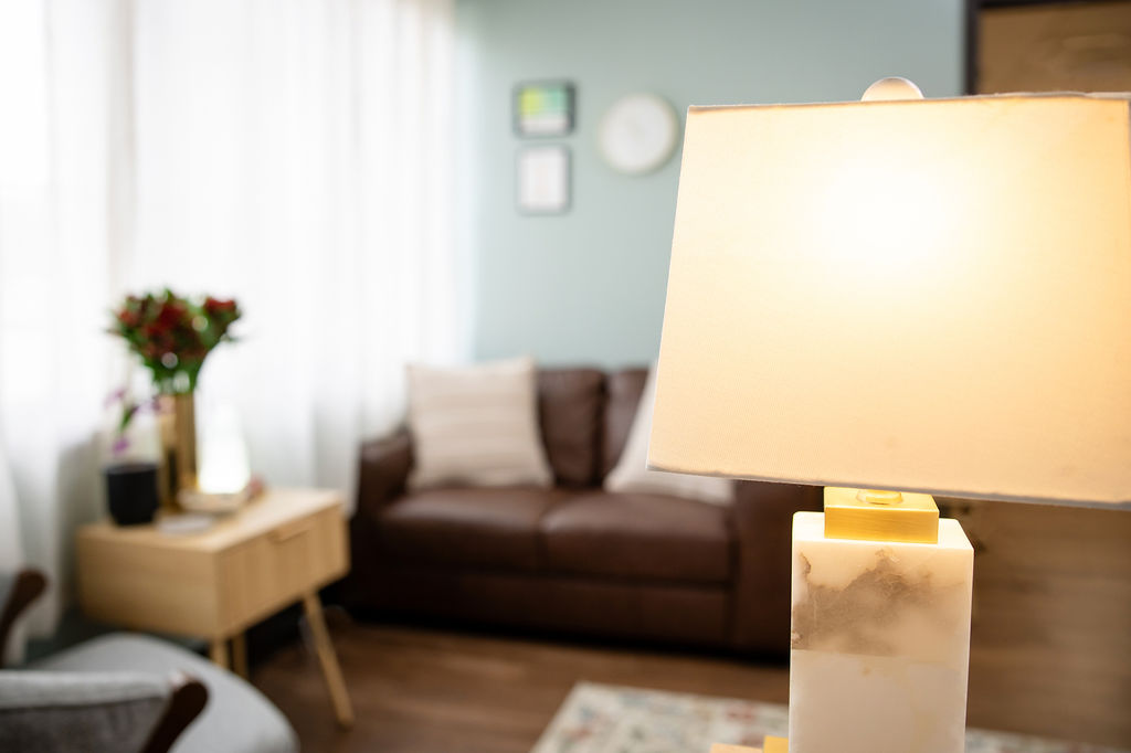 A lamp with brown leather couch and pillows in the background offering comfort in a well, but softly lit space. Sage walls and simple art tastefully decorate the space.
