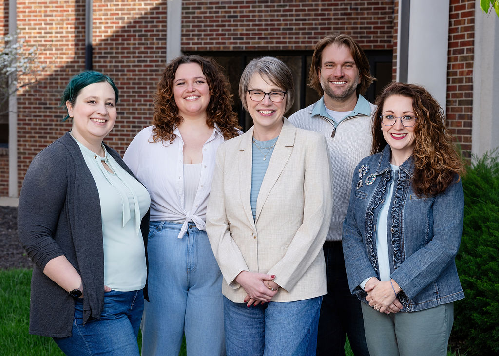 Group of 5 people, 4 feminine and one masculine, standing smiling outside with a brick wall as background.